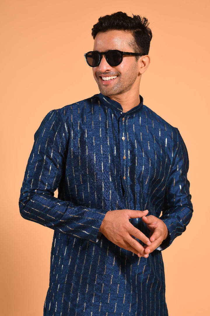 This Teal Blue silk-blend kurta is designed for comfort and elegance, featuring tone-on-tone threadwork with sequin detailing that runs horizontally for a striking yet subtle effect. Ideal for pujas, haldi, and festive occasions, the kurta is adorned with meenakari buttons along the front placket and finished with a mandarin collar. The knee-length hem adds to its traditional charm, making it a versatile choice for special celebrations.
